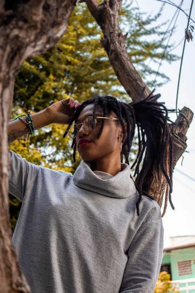 Modelo con dreadlocks y suéter gris junto a un árbol